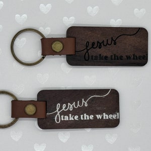May include: Two wooden keychains with brown leather straps and metal rings. Both keychains feature the text "Jesus take the wheel" in white and black lettering.