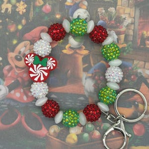 May include: A festive keychain featuring a Mickey Mouse-shaped candy cane design. The keychain is adorned with red, green, and white beaded accents. The design includes a silver clasp and key ring, perfect for holiday-themed accessories.