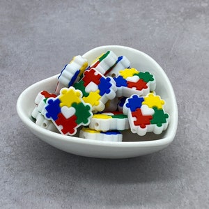May include: A bowl filled with colorful silicone puzzle piece beads with a white heart in the center of each piece. The beads are perfect for sensory play and can be used for necklaces, bracelets, or other crafts.