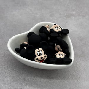 May include: A white heart-shaped bowl filled with black and white Mickey Mouse character charms. The charms are made of silicone and have a 3D design.