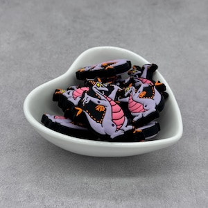 May include: A white heart-shaped dish filled with purple and pink dragon charms. The charms are made of rubber and have black outlines.