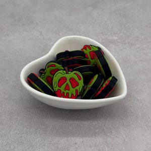 May include: A white heart-shaped bowl filled with green and red apple-shaped charms. The charms have a black border and a red skull design on the front. The bowl sits on a gray surface.