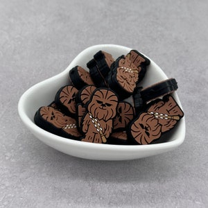 May include: A collection of brown and black silicone charms featuring a cartoon character with a furry face and a black headband. The charms are in a white heart-shaped bowl.