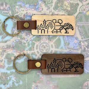 Può includere: Due portachiavi in legno con un cinturino in pelle marrone e un anello in metallo. Entrambi i portachiavi presentano un design inciso a laser dei monumenti iconici di Disney World: il Castello di Cenerentola, Spaceship Earth e la giostra 'it's a small world'.