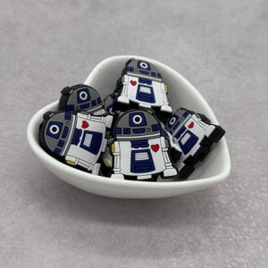 May include: A white bowl filled with small, blue, white and grey robot figurines with red hearts. The robots have a round head with a single eye and a small red heart on their chest.