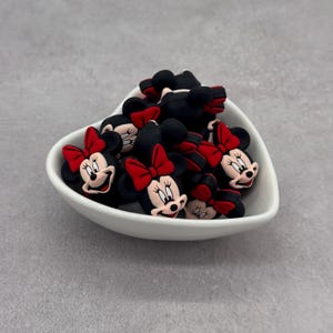 May include: A white heart-shaped bowl filled with black and red Minnie Mouse-themed charms. The charms feature Minnie's face, a red bow, and black ears. The bowl sits on a gray surface.
