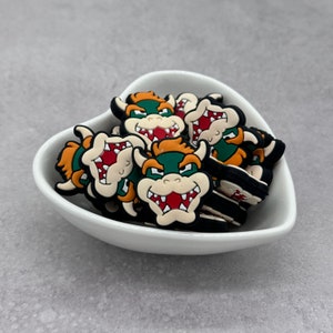 May include: A white ceramic bowl filled with multiple green, orange, and black cartoon character charms. The characters have large teeth and are smiling.