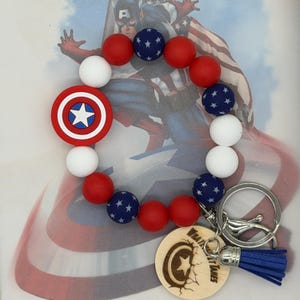 May include: A patriotic keychain bracelet featuring red, white, and blue beads. The bracelet includes a Captain America shield charm and a wooden tag with the text "WHAT IT TAKES". A blue tassel hangs from the keychain.