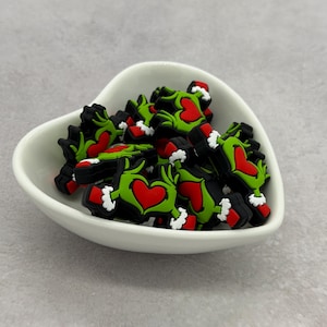 Pode incluir: Uma coleção de pequenos pingentes de borracha verdes, vermelhos e brancos em forma de mãos formando um coração, com punhos brancos nos pulsos. Estes pingentes decorativos estão contidos numa tigela de cerâmica branca em forma de coração.
