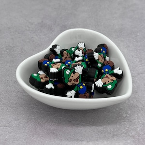 May include: A white ceramic bowl filled with green and black cartoon character charms. The charms are of a video game character wearing a green hat and overalls.