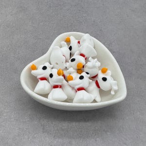 May include: A white heart-shaped bowl filled with small, white dog figurines. Each figurine has black eyes, a red collar, and an orange pumpkin nose. The figurines are a whimsical, cartoonish design.