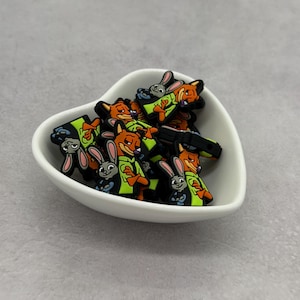 May include: A collection of cartoon character charms featuring a fox and a rabbit, in a white heart-shaped bowl. The fox is orange with a green jacket, and the rabbit is gray. The charms are black and colorful.