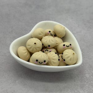 May include: A collection of small, cream-colored, round silicone beads shaped like steamed buns, each featuring a printed smiling face with black eyes, a curved mouth, and pink cheeks, contained within a white heart-shaped ceramic bowl.