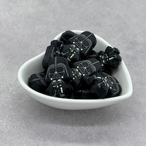 May include: A white heart-shaped bowl filled with black and green Star Wars Darth Vader character charms.