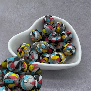 May include: A heart-shaped white bowl filled with colorful round beads. The beads have a geometric pattern in black, white, red, yellow, and blue.
