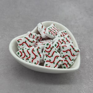 May include: A heart-shaped white ceramic bowl filled with white Christmas tree-shaped beads. The beads are decorated with red and green lines, resembling Christmas lights. The bowl sits on a gray surface.