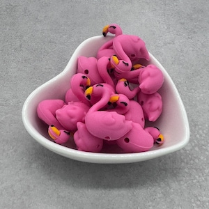 May include: A pile of pink flamingo-shaped silicone beads with black eyes and yellow beaks. The beads are in a white heart-shaped bowl.