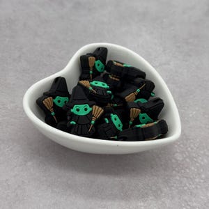 Może przedstawiać: Biała ceramiczna miska w kształcie serca wypełniona zielonymi i czarnymi figurkami czarownic. Figurki mają czarne kapelusze i zielone twarze. Każda czarownica trzyma miotłę.