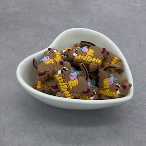 Puede incluir: Un grupo de dijes de ratón de dibujos animados marrones, amarillos y morados en un tazón blanco en forma de corazón. Los dijes están hechos de silicona y tienen un diseño 3D.