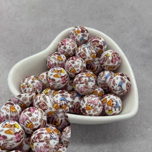 Peut inclure: Des perles rondes avec des motifs de personnages de dessins animés en rose, marron, jaune et bleu sur une base blanche. Les perles sont présentées dans un bol blanc en forme de cœur.