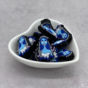 Puede incluir: Un bol blanco en forma de corazón lleno de abalorios de silicona azul y negro con un personaje de novia de dibujos animados. Los abalorios están diseñados para ser utilizados en la elaboración de joyas u otros proyectos de artesanía.