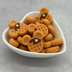 May include: A collection of orange and brown Mickey Mouse-shaped waffle beads in a white heart-shaped bowl. The waffles have a grid pattern and are topped with brown syrup and a square of butter. The beads are made of a smooth material.