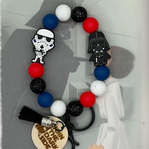 May include: A beaded keychain featuring Star Wars characters. The keychain includes red, white, black, and blue beads, along with charms of a Stormtrooper and Darth Vader. A wooden tag with the text "Join the Dark Side" is attached.