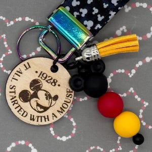 May include: A wooden keychain with a Mickey Mouse silhouette and the text "It All Started With A Mouse. 1928." The keychain also features a black and white Mickey Mouse patterned fabric strap, a yellow tassel, and black, red, and yellow beads.