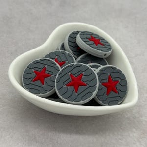 Peut inclure: Un ensemble de perles en silicone grises et rouges en forme d'étoile dans un plat blanc en forme de cœur. Les perles ont un fond gris avec une étoile rouge au centre. Le plat est posé sur une surface gris clair.