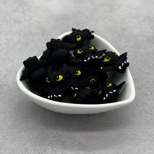 May include: A white heart-shaped bowl filled with small, black dragon figurines. Each dragon has bright yellow eyes and white accents on its wings and feet. The figurines are made of a smooth, matte material.