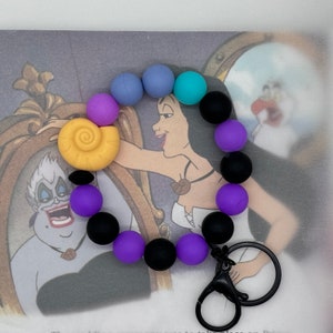 May include: A purple, blue, and black silicone bracelet with a seashell charm. The bracelet has a key chain attached to it.