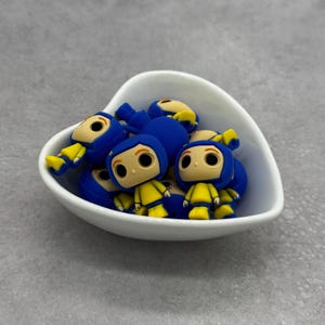 May include: A white heart-shaped bowl filled with blue and yellow cartoon character charms. The characters have blue hooded jackets, yellow raincoats, and brown eyes.