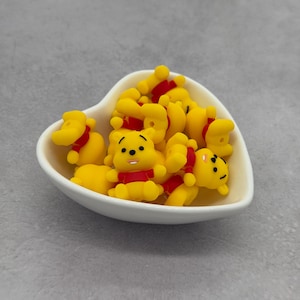 Puede incluir: Un cuenco blanco en forma de corazón lleno de figuras amarillas de Winnie the Pooh con camisas rojas. Las figuras tienen ojos negros y están en varias poses. El cuenco está sobre una superficie gris.