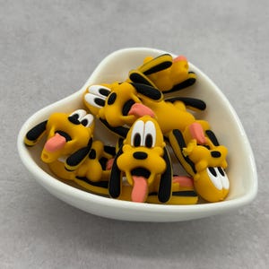 May include: A collection of yellow, black, and pink Pluto character charms in a white heart-shaped dish. The charms feature Pluto's signature black ears, pink tongue, and white eyes. The charms are made of a glossy material.