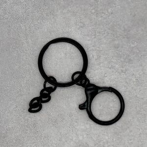 Può includere: Hardware per portachiavi in metallo nero con un portachiavi rotondo, una piccola catena e un moschettone. L'hardware è progettato per attaccare chiavi o piccoli accessori. Il portachiavi è mostrato su uno sfondo grigio chiaro.
