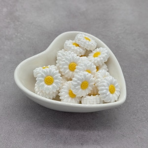 May include: A white heart-shaped bowl filled with white daisy-shaped beads with yellow centers. The beads are for crafting and jewelry making.