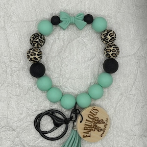 Op de afbeelding: Een ronde sleutelhangerarmband met mintgroene en zwarte ronde kralen, kralen met luipaardprint en een mintgroene strikbedel. Het bevat een zwarte metalen sluiting, een lichthouten ronde hanger met de tekst FABULOUS en een kwastje.