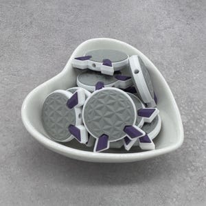 May include: A collection of gray and white round beads with a geometric pattern, accented with purple details, are displayed in a white heart-shaped dish. The beads have a hole for stringing and are suitable for jewelry making or crafts.