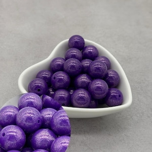 May include: A collection of round, purple beads with a glittery finish, displayed in a white heart-shaped bowl. The beads have a hole for stringing and are suitable for jewelry making or crafts.