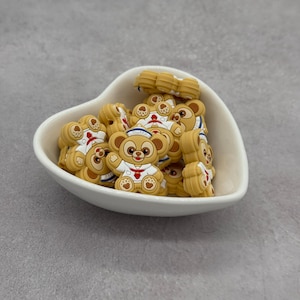 May include: A heart-shaped white ceramic bowl filled with numerous tan bear-shaped charms. Each charm features a bear wearing a white sailor outfit with a red tie and a blue hat. The charms are clustered together.