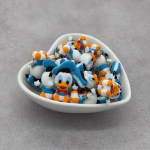 May include: A white heart-shaped bowl filled with small Donald Duck figurines. The figurines are white, blue, orange, and yellow, with a cartoonish design. The bowl sits on a gray surface.