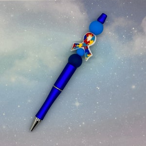 May include: A blue pen with a blue, red, yellow, and white puzzle piece ribbon charm. The pen is decorated with blue and white beads.
