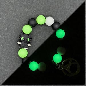 Peut inclure: Un bracelet de perles noir et vert avec un charm de dragon de dessin animé. Le bracelet comprend des perles noires, blanches et vert fluo. Certaines perles brillent dans le noir. Un porte-clés argenté est attaché.