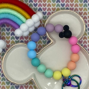 Puede incluir: Un llavero de cuentas de silicona arcoíris con un dije de Mickey Mouse negro y un llavero plateado. El llavero está en un plato blanco con forma de cabeza de Mickey Mouse.