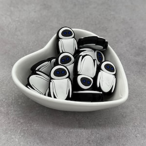 May include: A white heart-shaped bowl filled with black and white robot-shaped charms. The charms have blue eyes and are made of a glossy material. The bowl sits on a gray surface.
