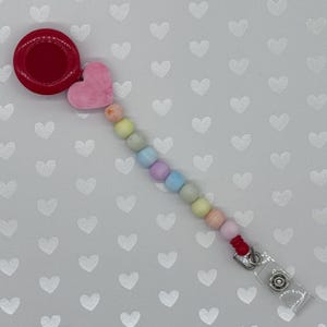 May include: A badge reel with a red circular top and a pink heart charm. A strand of pastel-colored beads connects to a clear plastic clip. The reel is designed to hold an ID badge or similar item.