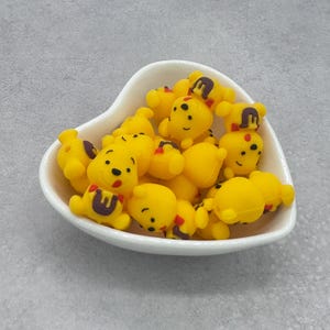 May include: A white heart-shaped bowl filled with yellow silicone Winnie the Pooh bear charms. The charms have black eyes, red noses, and brown details.