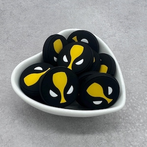 Pode incluir: Uma tigela branca em forma de coração cheia de pingentes redondos de silicone preto e amarelo com um rosto estilizado com olhos brancos e uma boca amarela.