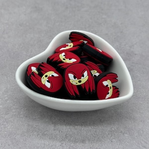 May include: A white heart-shaped bowl filled with red and black silicone charms featuring the face of a red hedgehog character.
