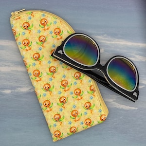 May include: A yellow eyeglass case with a zipper closure and a pattern of orange bird characters and blue flowers. Black sunglasses with rainbow lenses rest next to the case. The sunglasses have a black frame.